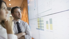 Two people standing up and looking at a large screen with graphs and numbers on it
