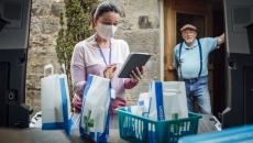 A woman using a tablet delivering medications while a man stands in his doorway in the background