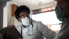 Healthcare provider wearing a mask, a lab coat and a stethoscope around their neck talking to a patient in their home