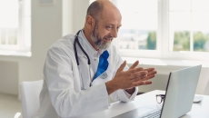 Healthcare provider sitting at a desk wearing a lab coat and a stethoscope while speaking to someone on a computer