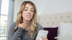 A woman rubbing her sore throat holding a smartphone.