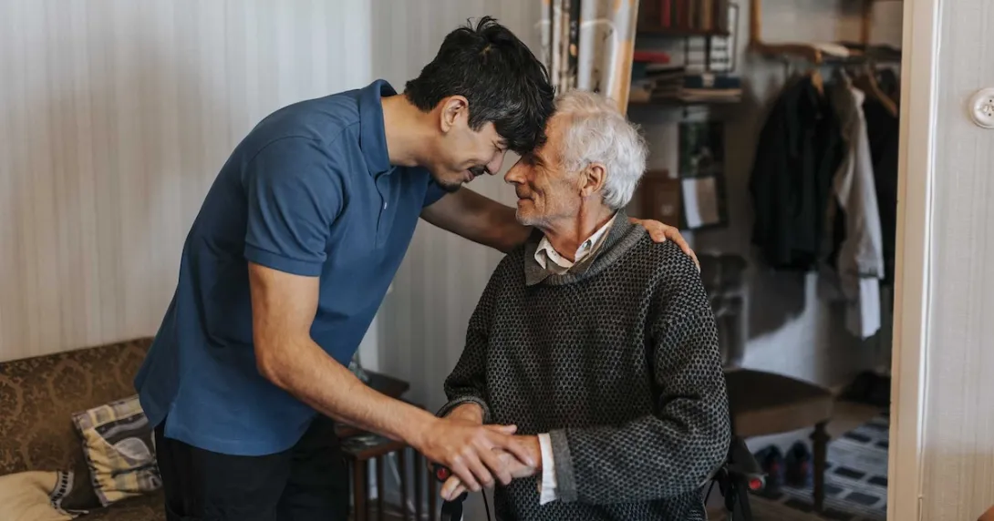 Two individuals with one in a wheelchair and the other standing over them with the two putting their heads together