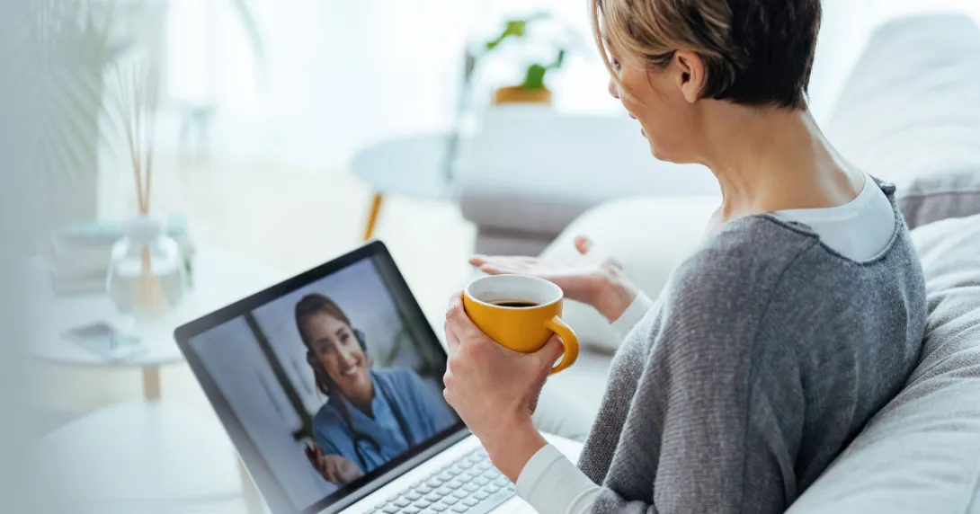 Person sitting on a couch holding a coffee cup while talking to a healthcare professional via a laptop