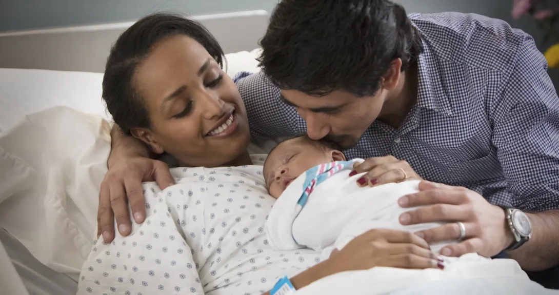 A person laying in a hospital bed with a newborn on their lap and another person standing beside them