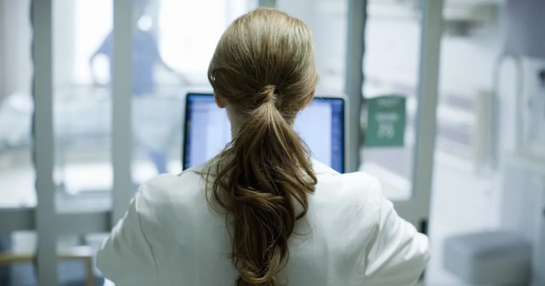 Woman doctor with long red hair using laptop in hospital, photographed from behind