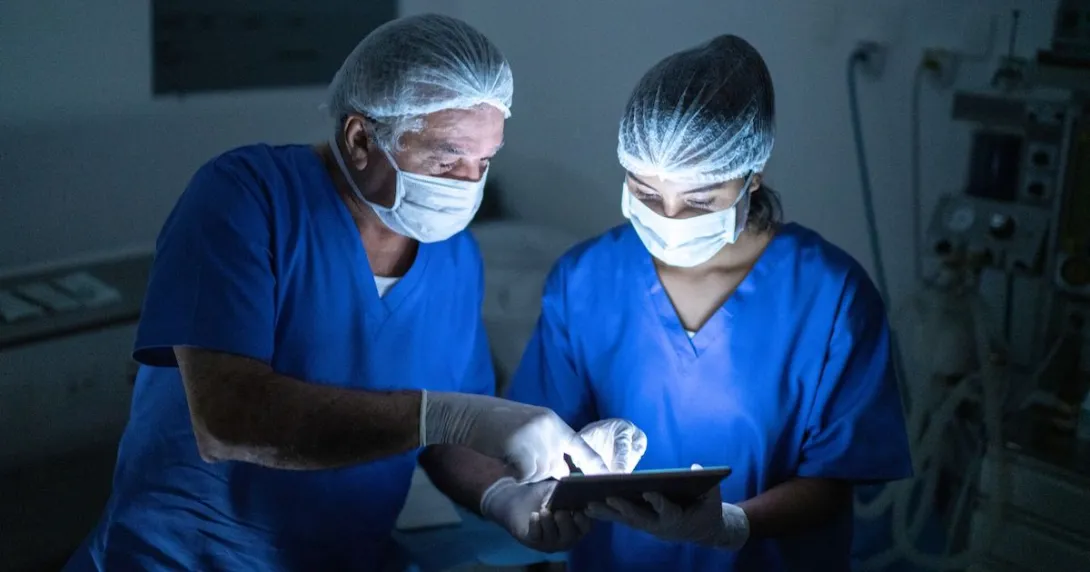 Two people wearing scrubs and masks in a procedure room look at a tablet.