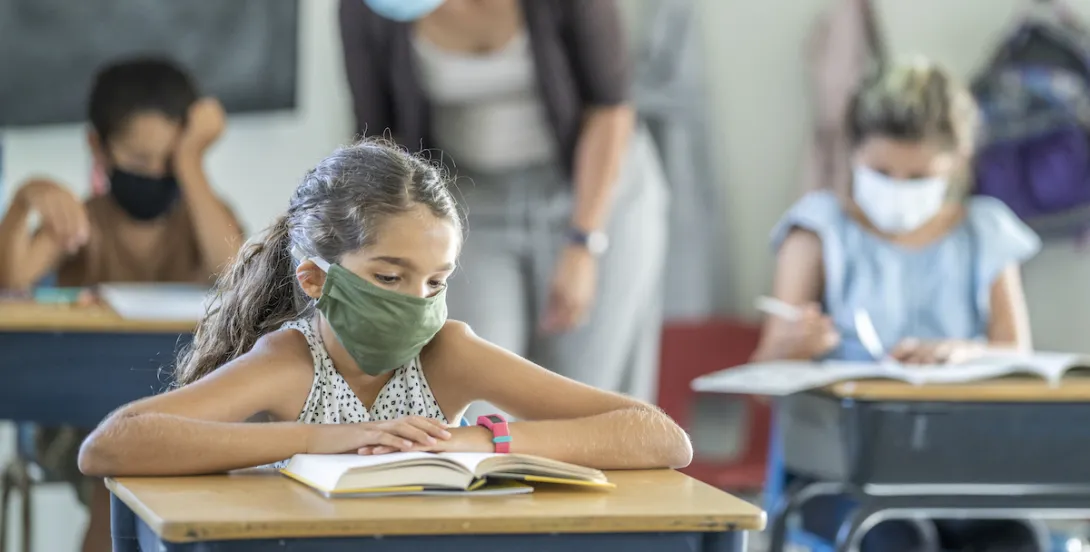 12 year old girl wearing a reusable, protective face mask in classroom while working on school work at her desk.