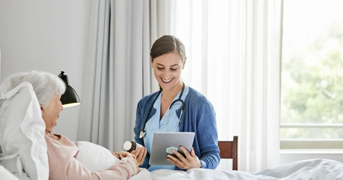 Person lying in bed with a healthcare provider sitting beside them in a chair holding a tablet