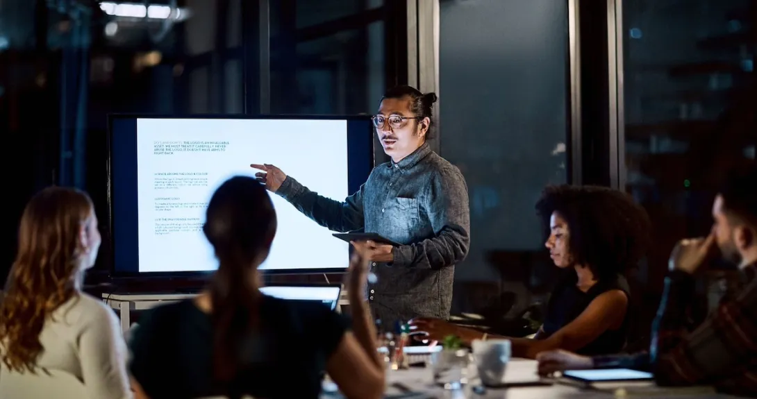 Person presenting in front of other people while standing next to a screen in a dark room