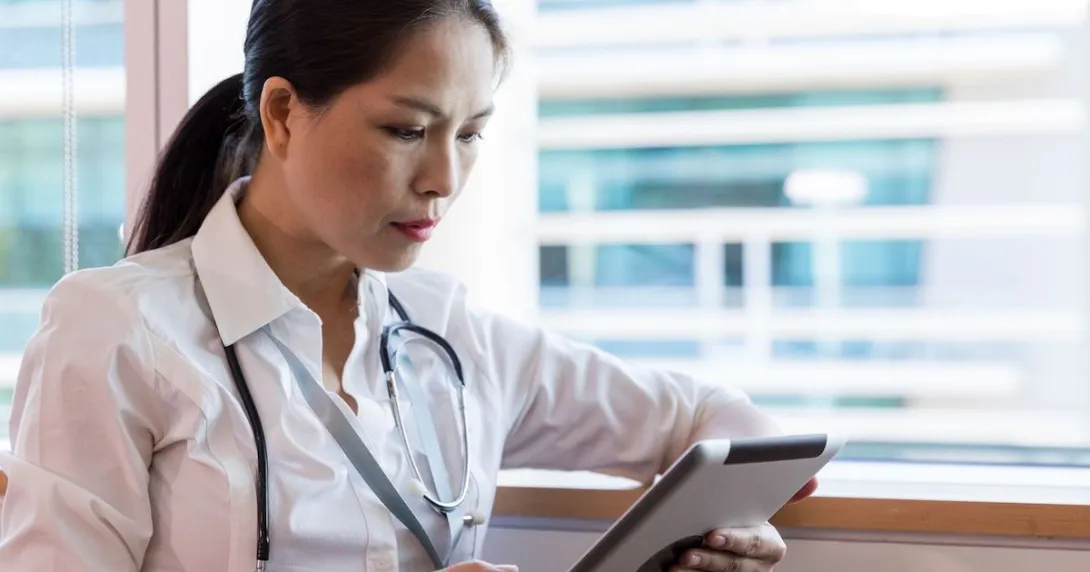 Healthcare provider sitting next to a window wearing a stethoscope around their neck and looking at a tablet