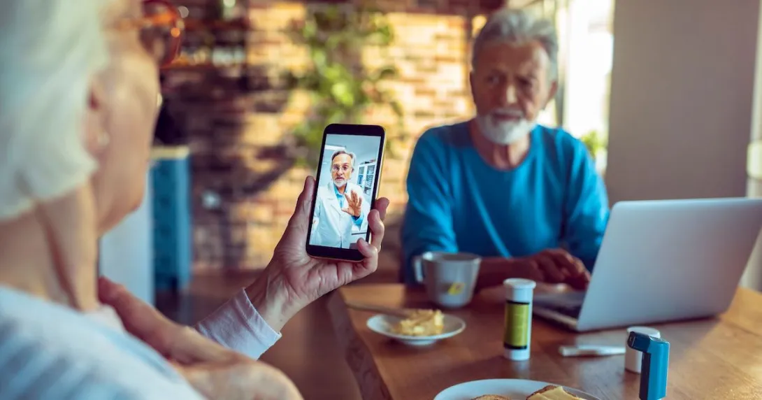 Two people sitting at a table with a computer on it and one of them is holding a phone