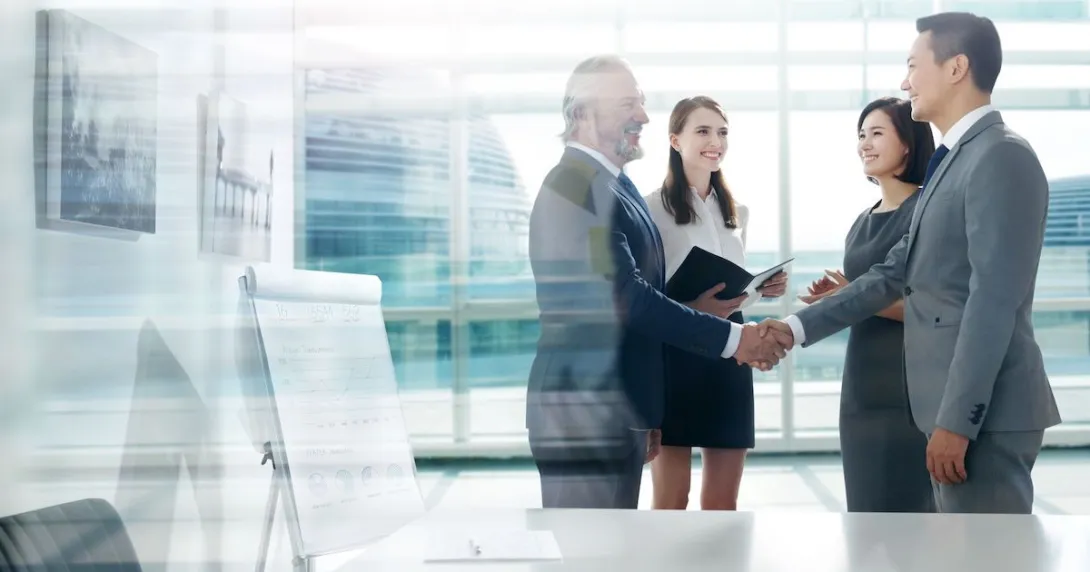 Four people standing in a room next to a large glass window with two of them shaking hands and the other two watching