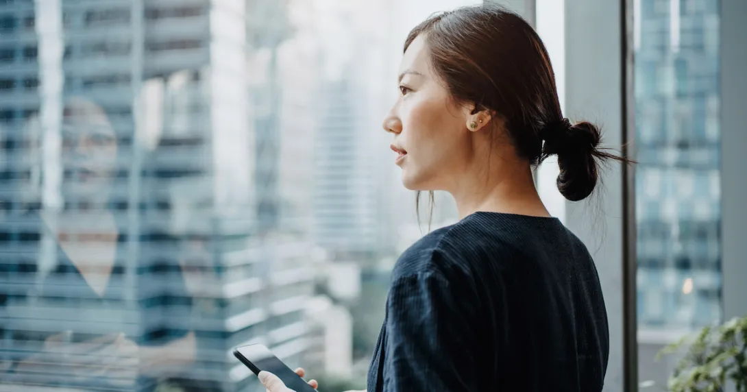 A young woman holding a smartphone and looking out a window.