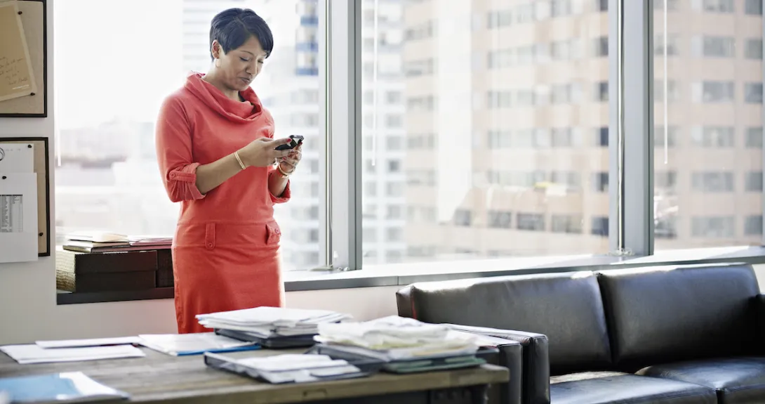 A woman standing at her desk using a smartphone.